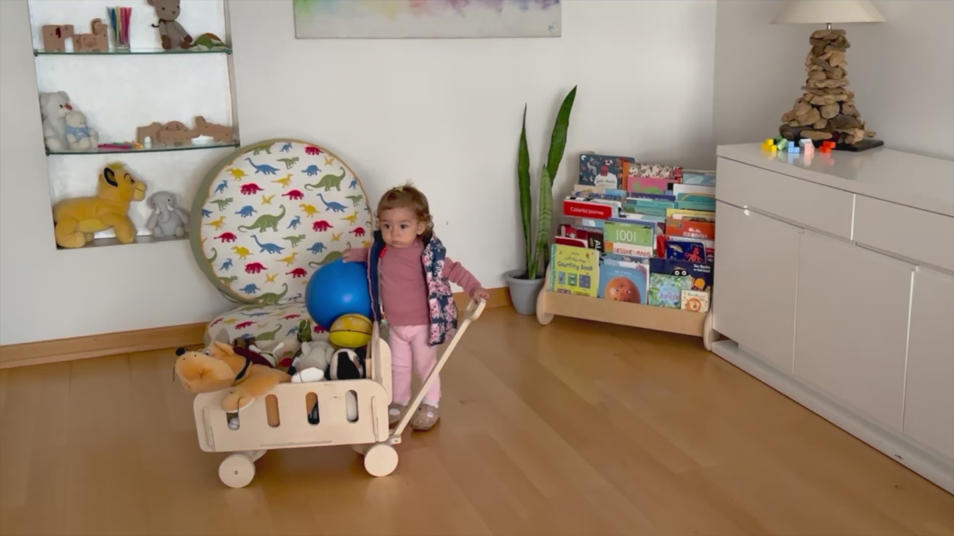 Toddler pushing eco-friendly wooden toy cart filled with colorful toys in a bright playroom.