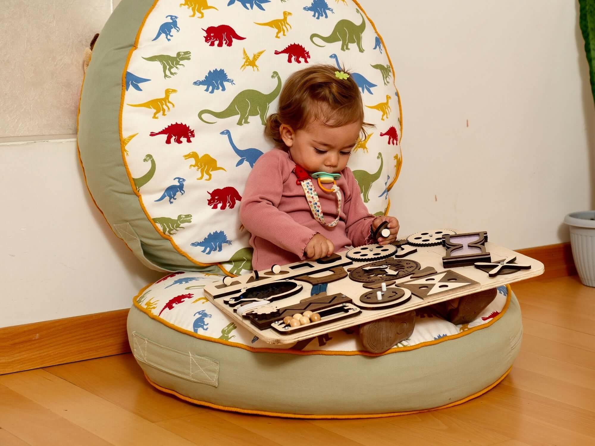 Toddler exploring a Wooden Busy Board with sensory activities while sitting on a colorful dinosaur bean bag.