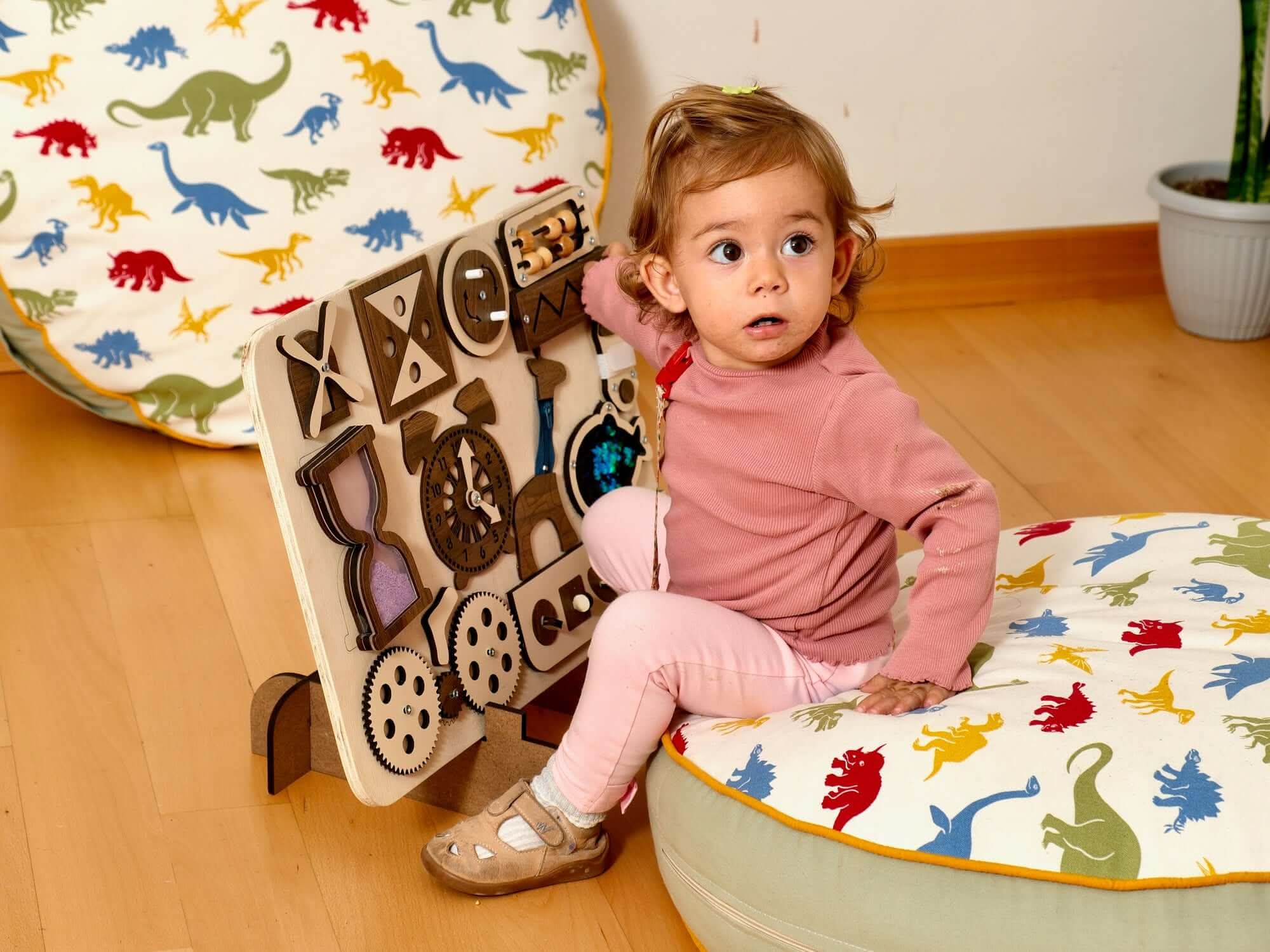 Toddler engaging with a Montessori-inspired wooden busy board and colorful dinosaur cushions.